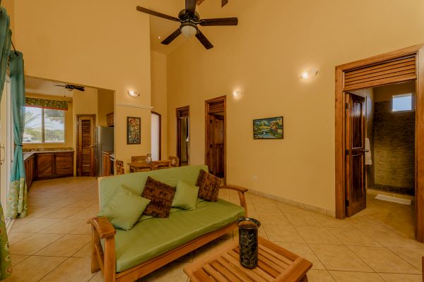 A cozy living room with wooden furniture, a green sofa, ceiling fan, art on the walls, and doors leading to other rooms. Includes a kitchen area.