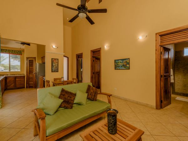 A cozy living room with wooden furniture, a green sofa, ceiling fan, art on the walls, and doors leading to other rooms. Includes a kitchen area.
