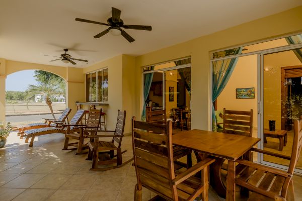 A covered patio with wooden chairs, dining tables, floor fans, and sliding doors leading inside. The outdoor seating area overlooks a lawn.