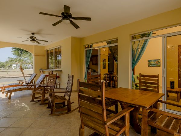 A covered patio with wooden chairs, dining tables, floor fans, and sliding doors leading inside. The outdoor seating area overlooks a lawn.