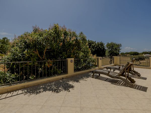 A sunlit patio with tiled flooring, lounge chairs, a garden with lush green trees and bushes, and a metal railing surrounding the area.
