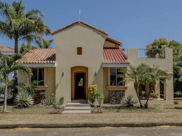 A beige stucco house with a red-tiled roof features a front entrance flanked by palm trees and desert plants, set against a clear blue sky.