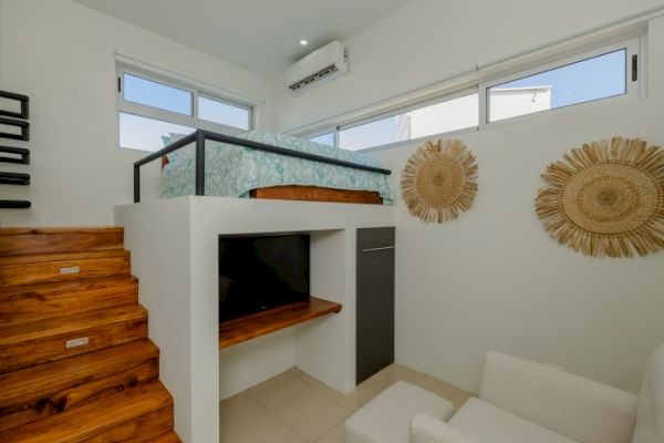 A small loft bedroom with a wooden staircase, bed on elevated platform, TV below the bed, white walls, large windows, and natural decor elements on the wall.