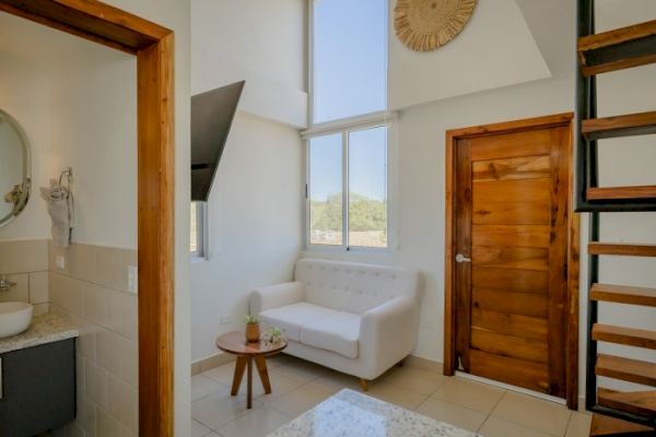 A modern, cozy room with a white couch, wooden side table, and staircase leading up. There's a bathroom to the left and a large window overlooking greenery.