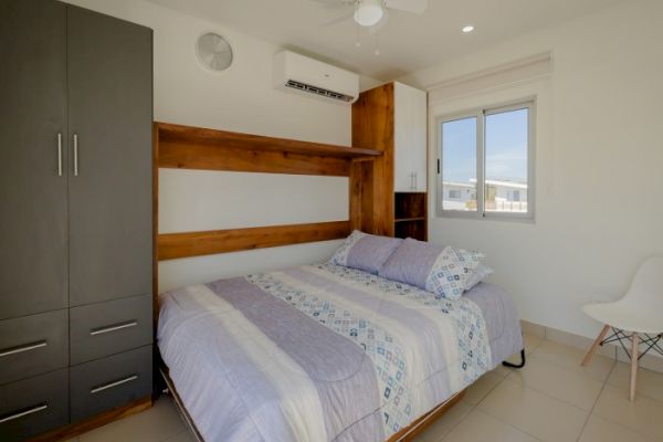 A bright bedroom featuring a neatly made bed, a wardrobe, a window, an air conditioner, a chair, and a ceiling fan in neutral hues.