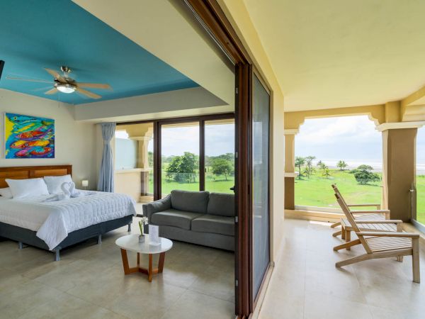 A spacious bedroom with a bed, sofa, and colorful painting opens up to a balcony with lounge chairs and a scenic view of greenery and the ocean.