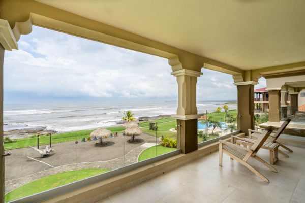 A beachfront view from a covered balcony with lounge chairs, overlooking a sandy beach, palm huts, and the ocean under a partly cloudy sky.