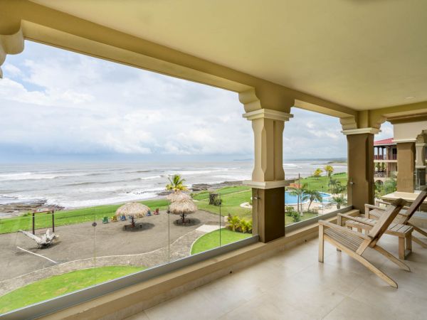 A beachfront view from a covered balcony with lounge chairs, overlooking a sandy beach, palm huts, and the ocean under a partly cloudy sky.