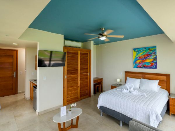 A modern bedroom with a blue ceiling, a large bed, a colorful painting above the headboard, a TV, and wooden furniture. White towels are on the bed.