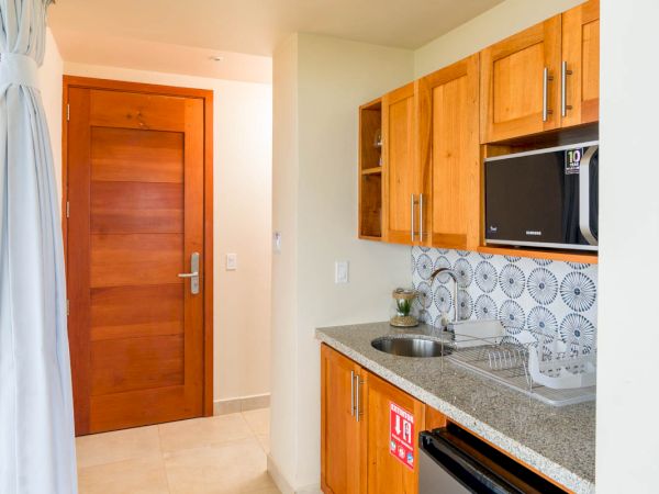 A compact kitchen with wooden cabinets, a small fridge, a microwave, a sink, and patterned backsplash tiles, leading to a wooden door.