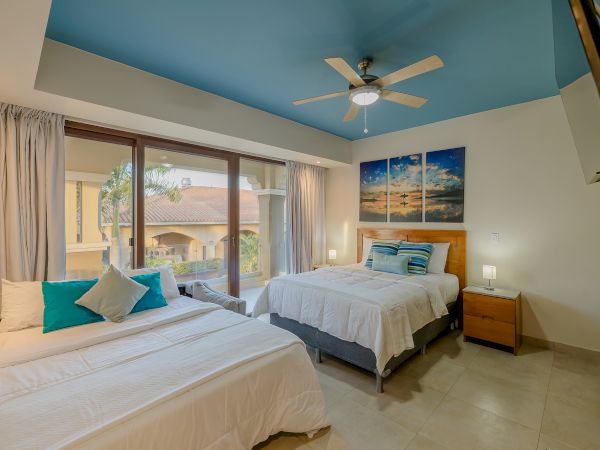 A bedroom with two beds, blue ceiling, wall art, a ceiling fan, bedside tables, lamps, and large windows with a view of a balcony and neighboring buildings.