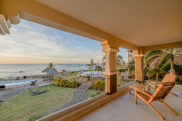 A beachfront patio with wooden chairs overlooks a scenic view of the ocean, a pool, palm trees, and a thatched-roof structure at sunset.