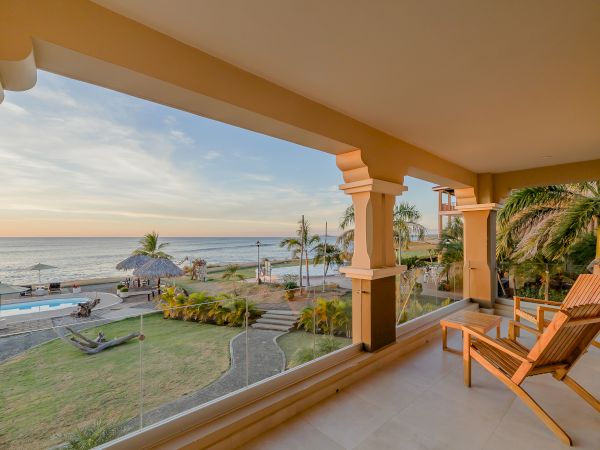A beachfront patio with wooden chairs overlooks a scenic view of the ocean, a pool, palm trees, and a thatched-roof structure at sunset.