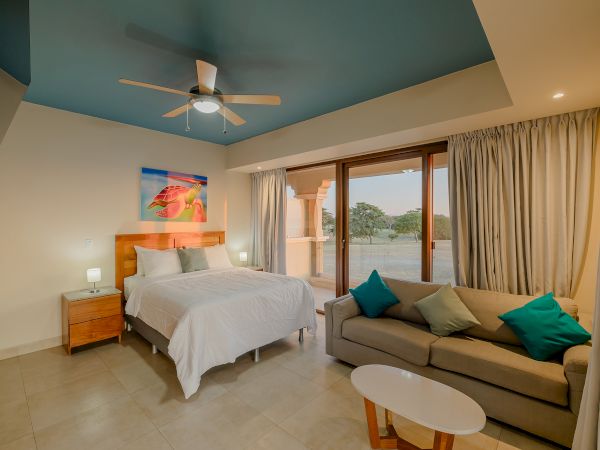 A cozy bedroom features a bed, sofa, nightstands, lamps, colorful pillows, wall art, ceiling fan, and a large window with a scenic view outside.