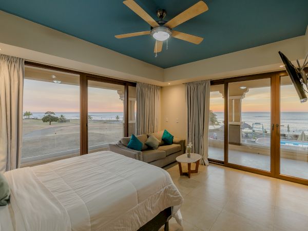 A bedroom with a double bed, sofa, ceiling fan, and wall-mounted TV, featuring large windows with an ocean view at sunset.