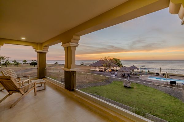 A beautiful beachside view from a covered patio, featuring wooden chairs, a pool, and a sunset over the ocean, with lush green grass and palm trees.