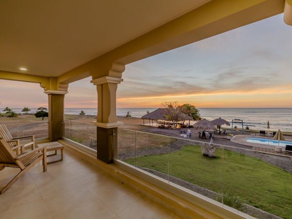 A beautiful beachside view from a covered patio, featuring wooden chairs, a pool, and a sunset over the ocean, with lush green grass and palm trees.