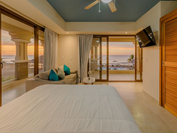 A cozy bedroom with a white bed, a small seating area, a wall-mounted TV, and large windows showcasing an ocean view at sunset.