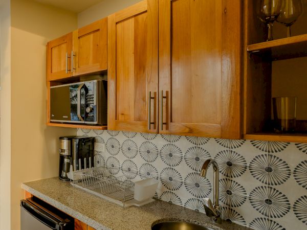 The image shows a small kitchen area with wooden cabinets, a granite countertop, a sink, a coffee maker, a microwave, and a tiled backsplash.