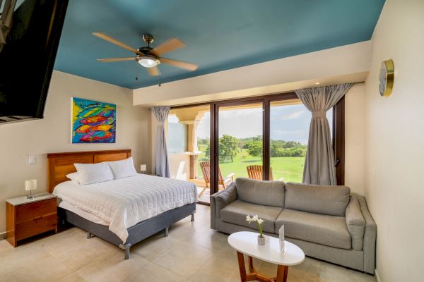 A bright, modern bedroom with a double bed, couch, and wall art. Sliding glass doors lead to a balcony with a scenic green view.