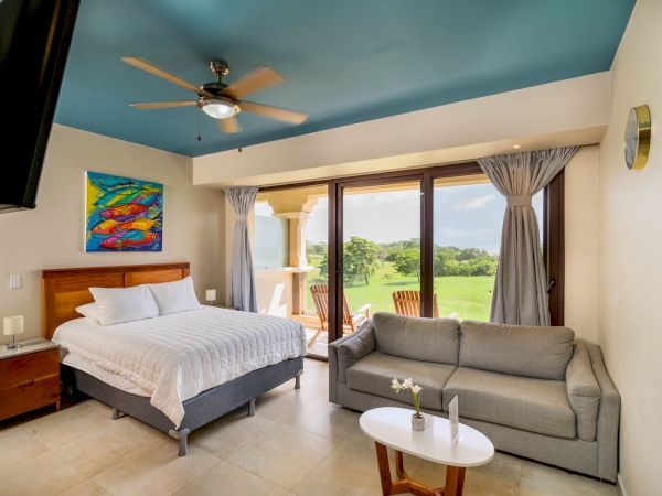 A bright, modern bedroom with a double bed, couch, and wall art. Sliding glass doors lead to a balcony with a scenic green view.