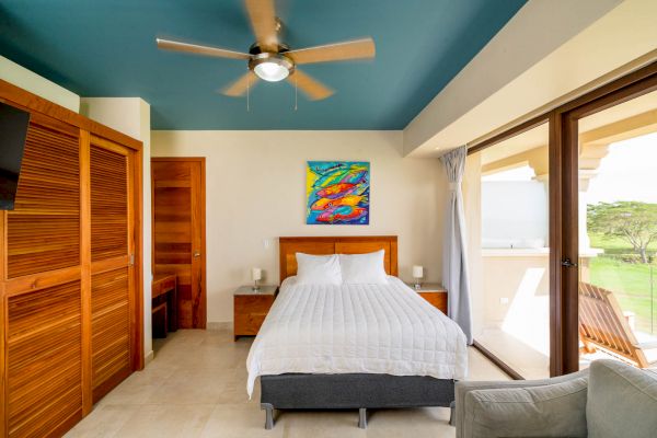 A modern bedroom with a blue ceiling, a bed, wooden furniture, a colorful painting, and large glass doors opening to a balcony with a scenic view.