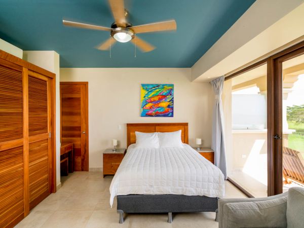 A modern bedroom with a blue ceiling, a bed, wooden furniture, a colorful painting, and large glass doors opening to a balcony with a scenic view.