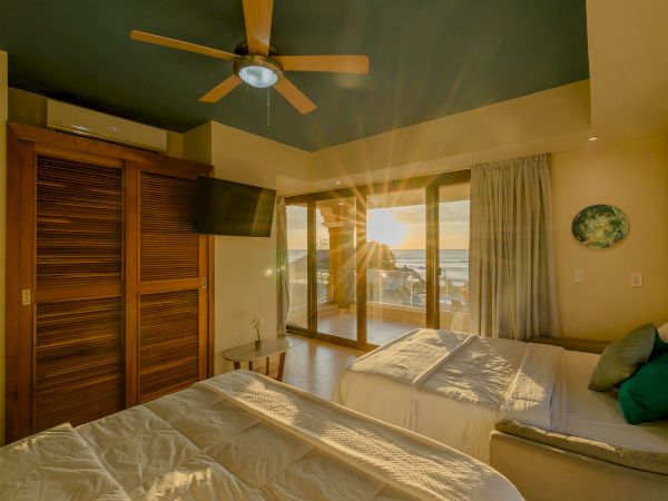 A cozy bedroom features two beds, a ceiling fan, a wall-mounted TV, and sliding doors opening to a sunset-lit balcony, with a beach view outside.
