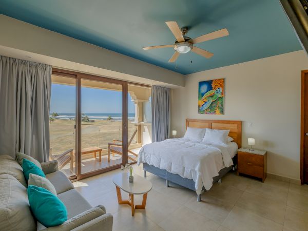 A cozy beachside bedroom with a queen bed, sofa, coffee table, ceiling fan, vibrant painting, and a stunning ocean view from large windows ending the sentence.
