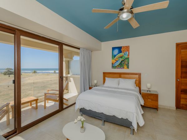 A beachside bedroom with a bed, nightstands, ceiling fan, and artwork on the wall. It has large windows and a door leading to a balcony.