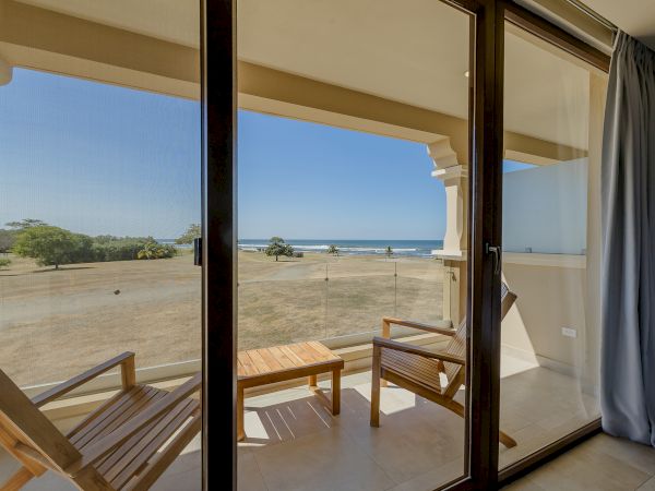The image shows a balcony with two wooden chairs and a table, overlooking a scenic beach with palm trees and clear skies, viewed through glass doors.
