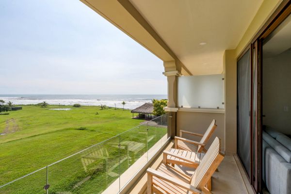 A balcony with wooden chairs overlooks a grassy area and a beach with the ocean in the background. Glass railing and sliding doors are visible.