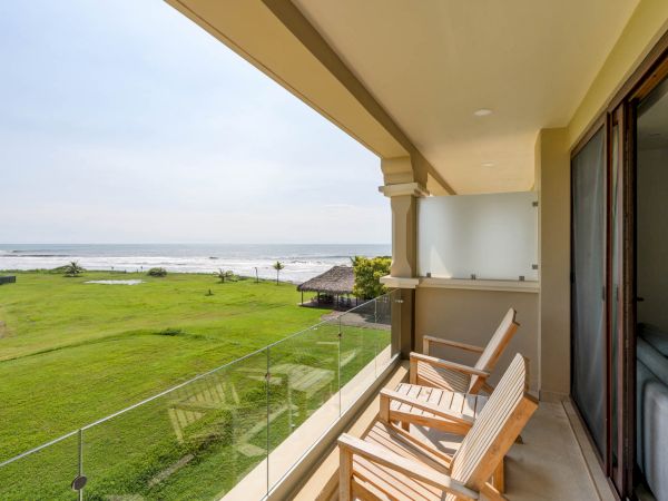 A balcony with wooden chairs overlooks a grassy area and a beach with the ocean in the background. Glass railing and sliding doors are visible.