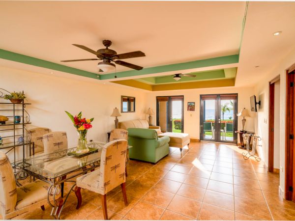This image shows a bright living and dining area with tiled floors, a ceiling fan, and a glass dining table. There's a view of the ocean through the doors.