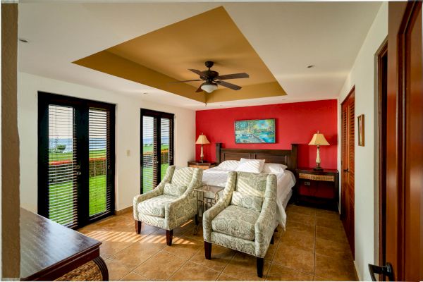 A cozy bedroom features a large bed, two armchairs, a ceiling fan, and a vibrant red accent wall with a painting. The room opens to a view outside.