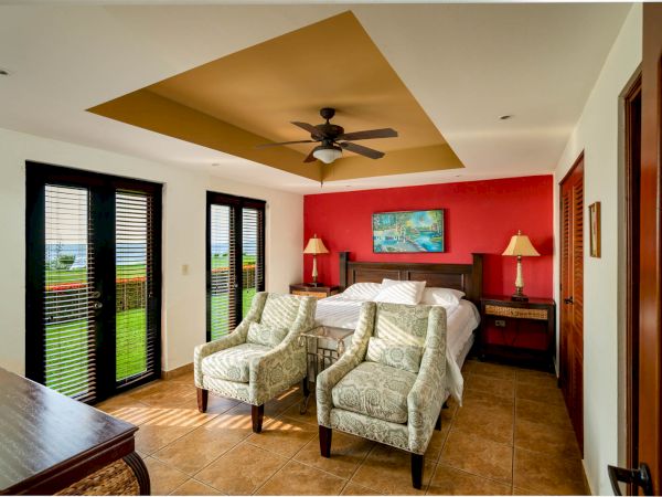 A cozy bedroom features a large bed, two armchairs, a ceiling fan, and a vibrant red accent wall with a painting. The room opens to a view outside.