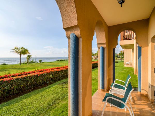 A seaside patio with arches, two chairs, and a serene view of a lush green lawn, palm trees, and the ocean in the distance.