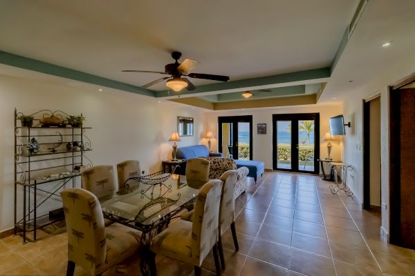 This image shows a cozy living-dining area with a glass table, upholstered chairs, a sofa, and a TV. The room has large windows with an ocean view.