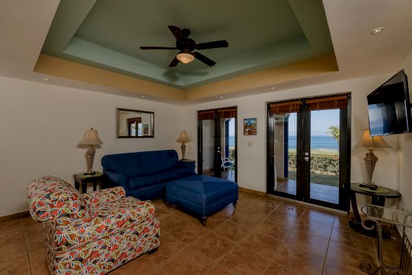 A cozy living room with a sofa, colorful armchair, coffee table, ceiling fan, and TV, and a view of the outside through glass doors.