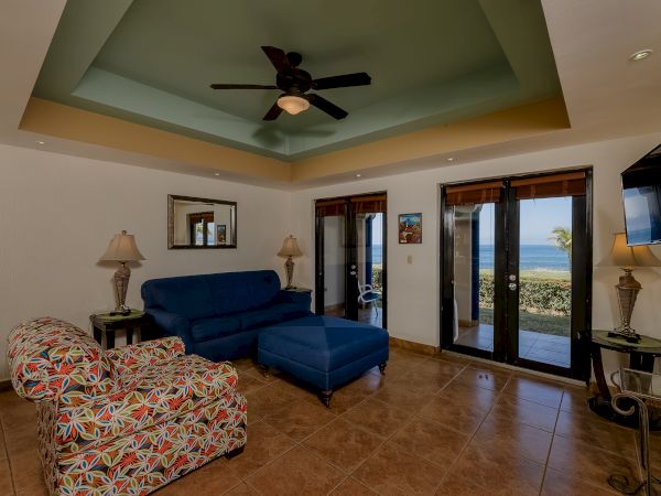A cozy living room with a sofa, colorful armchair, coffee table, ceiling fan, and TV, and a view of the outside through glass doors.