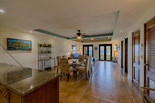 A spacious living room with tile floors, a dining area, and a kitchen countertop. There's a ceiling fan and large windows leading to a balcony.