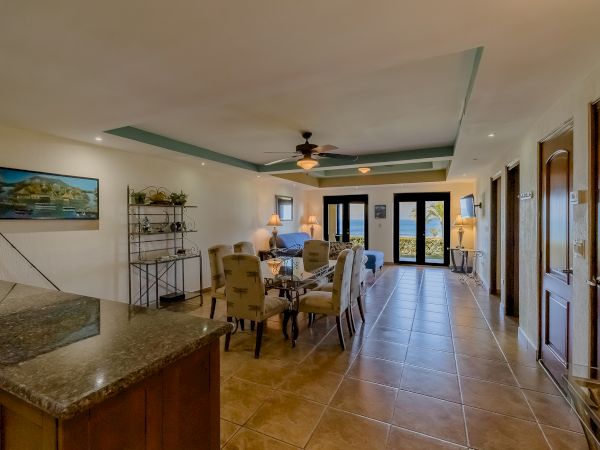 A spacious living room with tile floors, a dining area, and a kitchen countertop. There's a ceiling fan and large windows leading to a balcony.