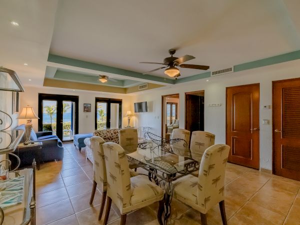 A well-furnished living and dining area with a ceiling fan, glass table, upholstered chairs, and glass doors opening to a balcony overlooking a scenic view.