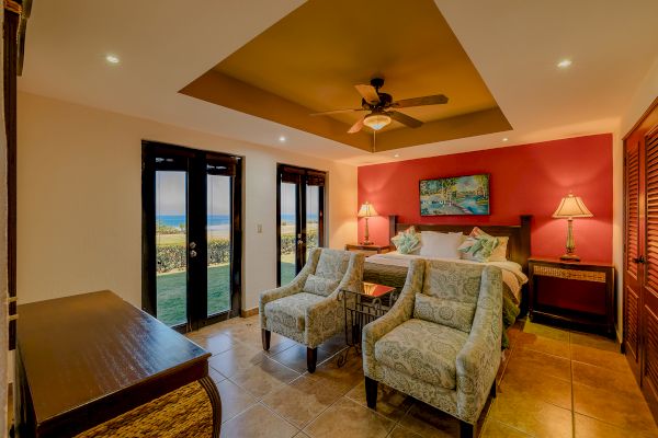 A cozy bedroom with a king-sized bed, armchairs, lamps, a ceiling fan, and a view of the outdoors through French doors.