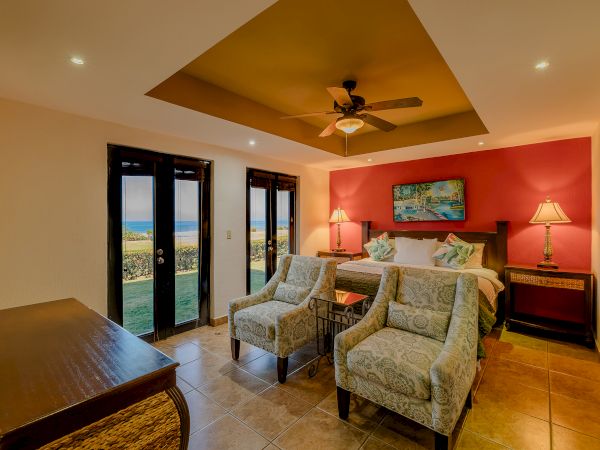 A cozy bedroom with a king-sized bed, armchairs, lamps, a ceiling fan, and a view of the outdoors through French doors.