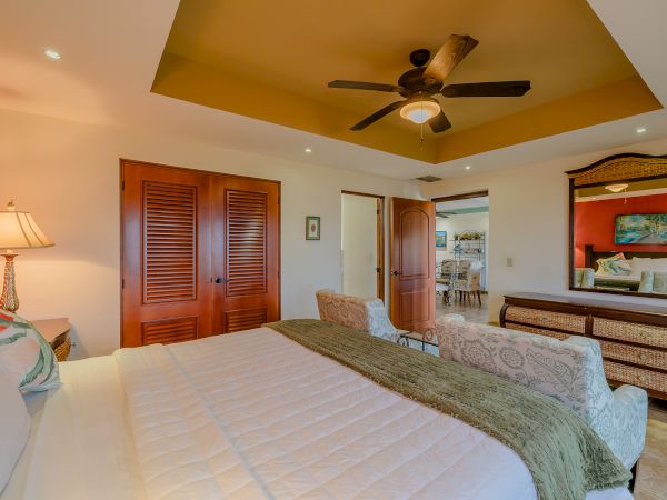 A cozy bedroom with a large bed, ceiling fan, wooden closet doors, mirrored dresser, and an open door leading to another room in the house.