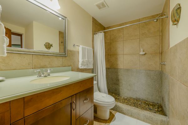 This image shows a bathroom with a vanity, rectangular mirror, toilet, towel rack, and a shower area with beige tiles and a white shower curtain.