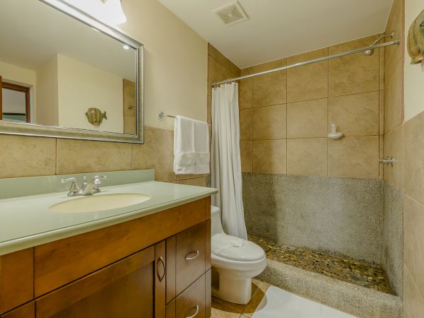 This image shows a bathroom with a vanity, rectangular mirror, toilet, towel rack, and a shower area with beige tiles and a white shower curtain.