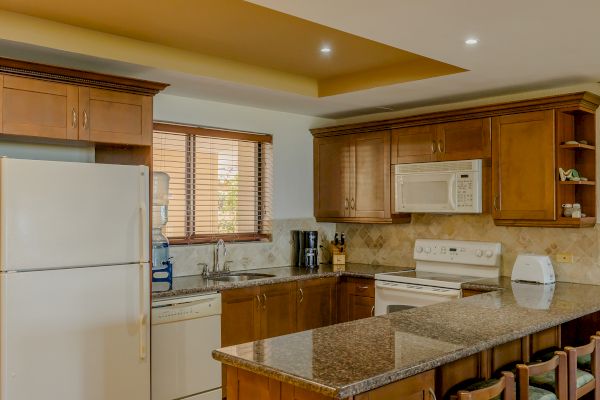 A kitchen with wooden cabinets, granite countertops, a white fridge, dishwasher, microwave, stove, and sink, with bar stools at the island.