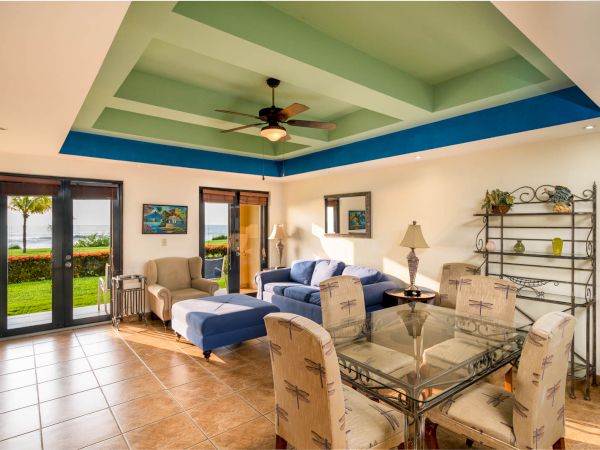 A spacious living room features a dining table, sofas, chairs, ceiling fan, decor, large windows, and doors opening to a garden with a scenic view.